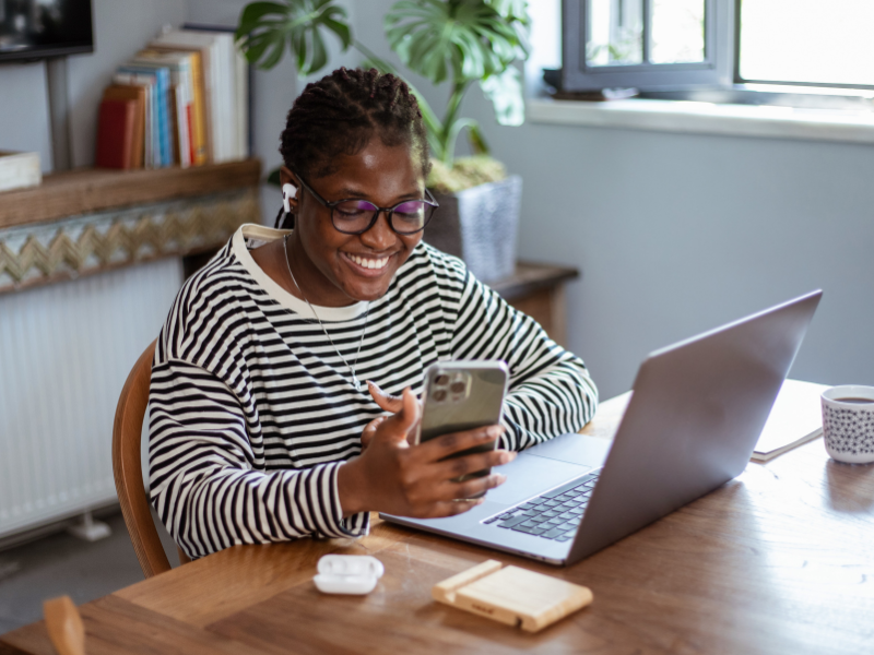 A woman sitting at a table looking at her phone in front of her laptop.