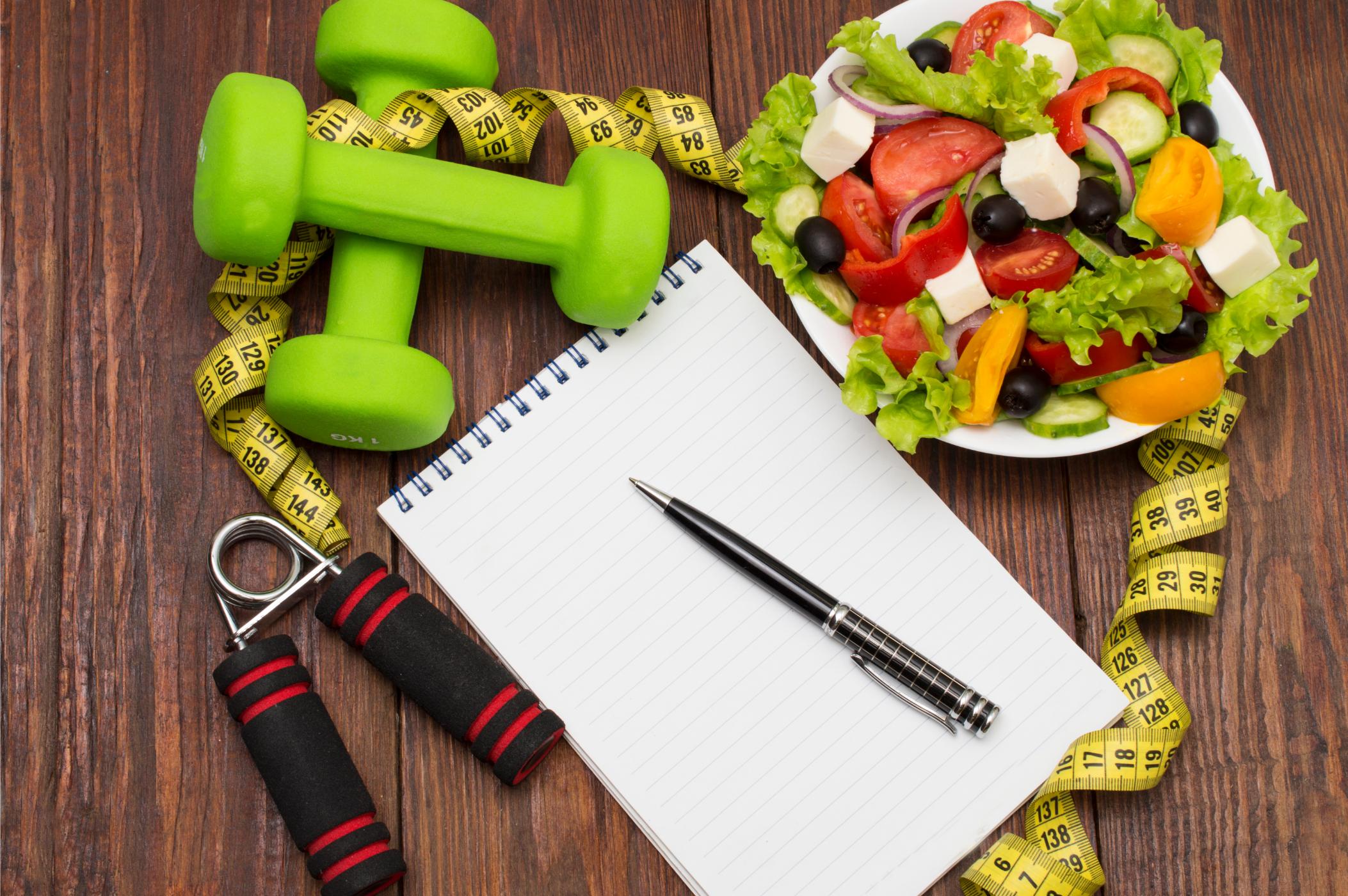 A notepad and pen surrounded by weights and healthy foods.