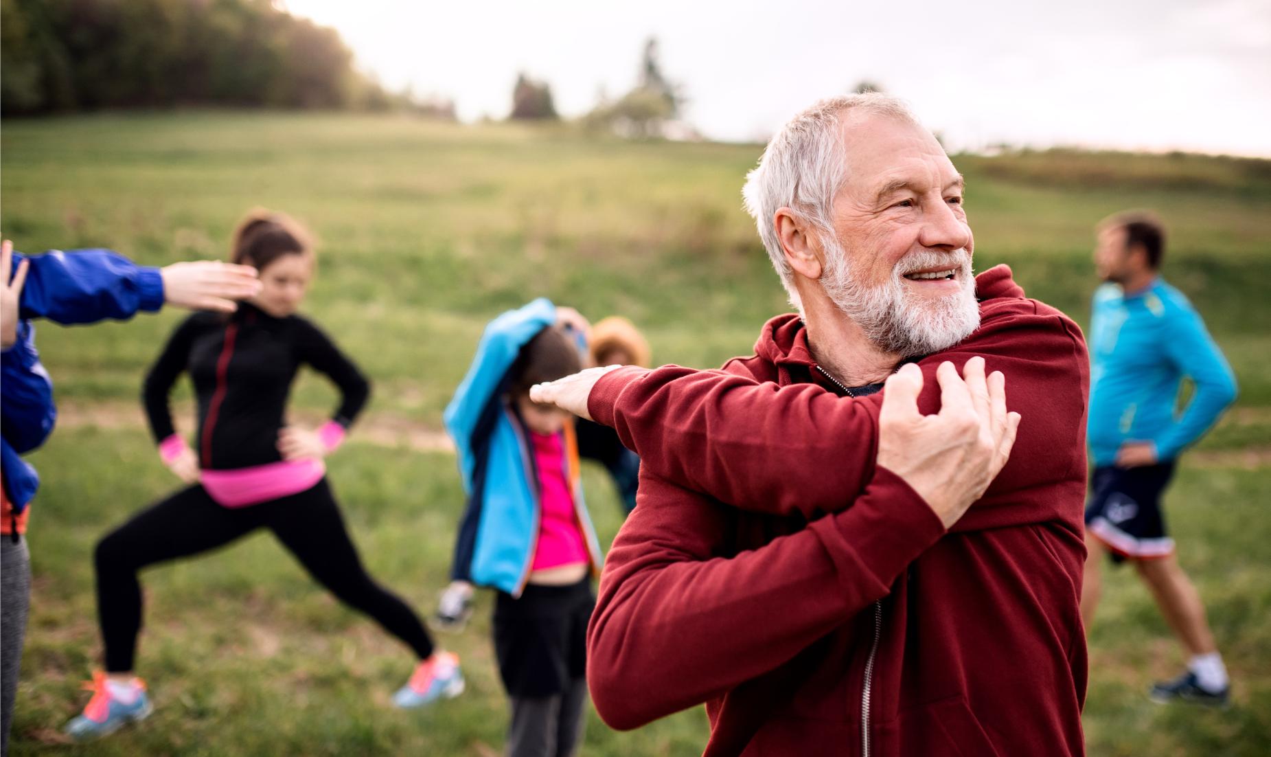 An older man stretching his arm outside in a group setting.